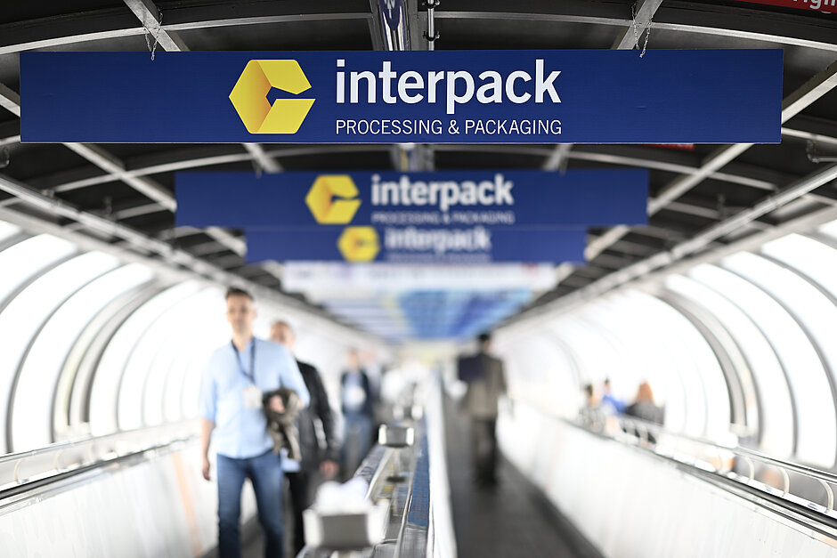 Copyright: Constanze Tillmann, Mintarder Weg 34, 40472 Duesseldorf, Germany Ljudi hodaju pokretnom stazom ispod nekoliko plavih znakova sa žutim logotipom Interpacka i tekstom 'Interpack OBRADA I PAKIRANJE' u strukturi nalik tunelu.